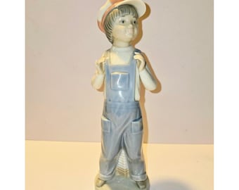 May include: A porcelain figurine of a young person wearing blue overalls and a white and orange cap. The figurine is holding a white cloth over their shoulders. The figure is standing on a round, light gray base.