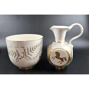 May include: A cream-colored ceramic bowl and pitcher set. The bowl features a gold fern design. The pitcher has a gold horse emblem and a handle. Both pieces have a crackle glaze finish.