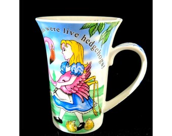 May include: White ceramic mug featuring a whimsical illustration from Alice in Wonderland. The design includes a girl, a flamingo, and the text "balls were live hedgehogs!" The mug has a curved handle and a flared rim.