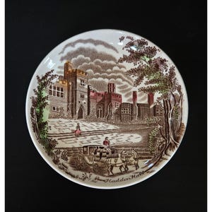 May include: A round ceramic saucer featuring a brown transferware print of Haddon Hall. The scene depicts a large stone manor, a horse-drawn carriage, and a person walking on the grounds. The text Haddon Hall is printed at the bottom.
