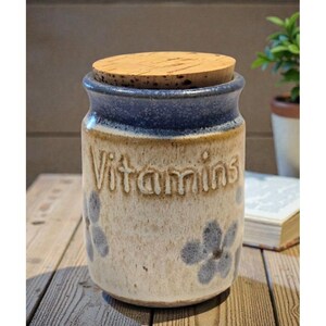 Könnte beinhalten: Ein Keramiktopf mit Korkdeckel, beschriftet mit "Vitamins". Der Topf hat eine beige Basis mit blauen Blumenakzenten und einem dunkelblauen Rand. Er steht auf einer Holzoberfläche, was eine rustikale oder ländliche Ästhetik suggeriert.
