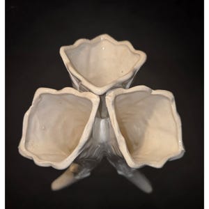 May include: A white ceramic vase with three flared openings, each with a scalloped edge. The vase has a textured, organic design and sits on three supporting legs. The background is black.