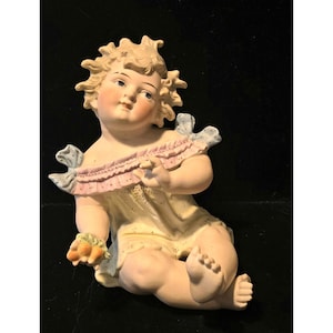 May include: A porcelain figurine of a seated cherub with blonde, spiky hair. The cherub wears a yellow dress with pink and blue accents. The figurine is detailed with delicate facial features and holds a small bunch of flowers.