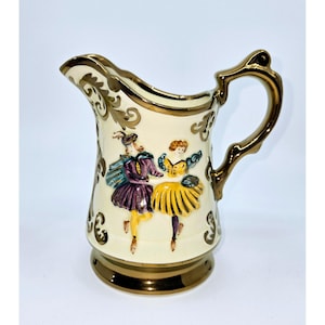 May include: A cream-colored ceramic pitcher with a gold-colored handle and trim. The pitcher features a raised design of two figures in colorful clothing dancing.