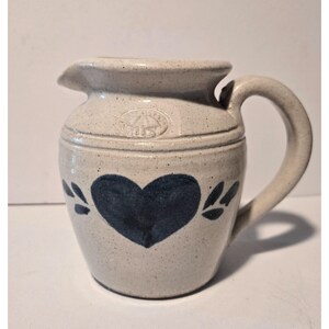 May include: A stoneware pitcher with a light beige speckled finish. The pitcher features a dark blue heart design on the front, with leaf accents. It has a curved handle and a spout for pouring. The pitcher has a rustic, handmade aesthetic.