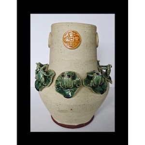 Puede incluir: Un jarrón de cerámica beige con base redonda y abertura ancha. El jarrón presenta tres diseños de ranas y hojas de nenúfar verdes, un emblema circular y dos asas laterales. El jarrón mide aproximadamente 30 cm de alto.