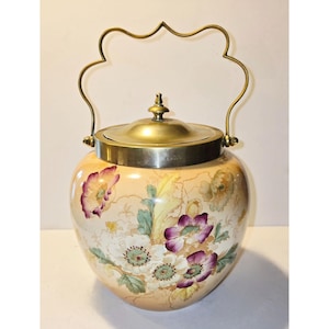 Puede incluir: Un tarro de galletas de cerámica vintage con un diseño floral. El tarro tiene una base de color melocotón con flores blancas y moradas pintadas. Cuenta con una tapa y un asa de latón, añadiendo un toque de elegancia a la decoración.