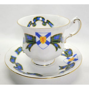 May include: A white teacup and saucer set with a blue, green and yellow tartan pattern. The cup features a white shield with a blue cross and a red lion. The saucer has a similar tartan pattern with a smaller shield.