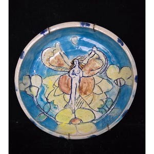 Può includere: Una ciotola in ceramica dipinta a mano con interno blu e bordo bianco. La ciotola presenta una figura centrale con ali di farfalla, circondata da fiori gialli e una luna crescente. L'opera è firmata.