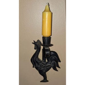 Puede incluir: Un portavelas de pared de metal negro con forma de gallo y una vela amarilla. El portavelas está montado en una pared y presenta un diseño detallado de gallo. La vela es cilíndrica y tiene una parte superior puntiaguda.