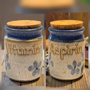 May include: Two ceramic canisters with cork lids, featuring a cream-colored base with blue rims and blue floral accents. One canister has the word Vitamins embossed on the front, and the other has the word Aspirin embossed on the front.