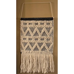 May include: A woven wall hanging with a geometric pattern in black and cream. The design features triangles and rows of small, raised dots. The top edge is black, and the bottom has long cream-colored fringe. A white metal frame and string are attached for hanging.