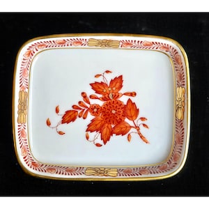 May include: A rectangular white ceramic dish with a floral design. The flowers are orange and red, with gold accents on the rim. The dish has a gold border and is likely for decorative use.