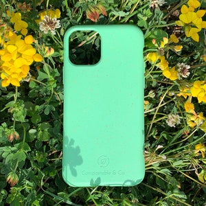 May include: A light green phone case with a speckled texture, set against a backdrop of green foliage and yellow flowers. The case has a cutout for the camera and the text "Compostable & Co." is visible.