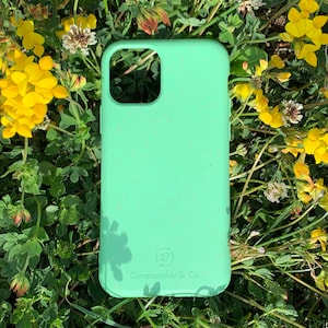 May include: A light green phone case with a speckled texture, set against a backdrop of green foliage and yellow flowers. The case has a camera cutout and the text "Compostable & Co." is visible.