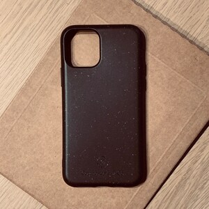 Puede incluir: Una funda de teléfono marrón oscuro con una textura moteada, sobre una superficie de papel marrón. La funda tiene un recorte para la cámara y las palabras "Compostable by Co." están impresas en la parte inferior. La funda está diseñada para proteger un teléfono.