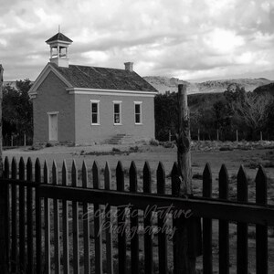 May include: Black and white photograph of a small, brick building with a bell tower, set against a cloudy sky. A wooden picket fence frames the foreground, with a natural landscape in the background.