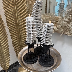 Peut inclure: Trois bougies piliers décoratives avec un motif texturé gris et blanc, chacune surmontée d'une lumière LED vacillante. Les bougies sont posées sur des supports en métal noir, disposées sur un plateau argenté. Le plateau repose sur une petite table à trois pieds.