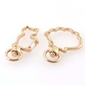 May include: Two gold-colored metal swivel clasps. One clasp is shaped like a cat, the other like a cloud. Each has a small ring at the bottom and a spring-loaded clasp mechanism. These clasps are likely used for keychains or jewelry.