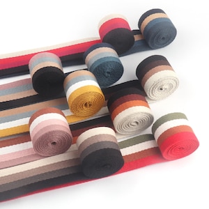 Può includere: Una collezione di rotoli di fettuccia a righe e fettuccia piatta. I rotoli mostrano varie combinazioni di colori, tra cui rosso, nero, blu e marrone. La fettuccia piatta presenta strisce abbinate, creando un design coerente per l'artigianato e gli accessori.