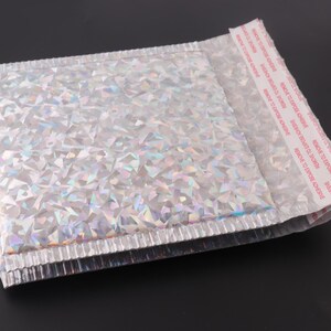 May include: A silver iridescent padded mailing envelope with a peel-and-seal adhesive strip. The adhesive strip has the text "REMOVE TO EXPOSE ADHESIVE" printed on it.