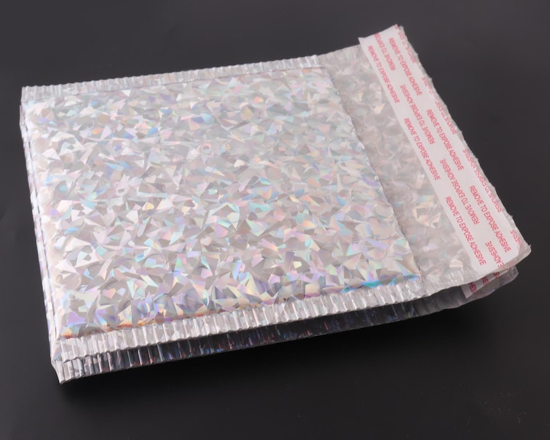 May include: A silver iridescent padded mailing envelope with a peel-and-seal adhesive strip. The adhesive strip has the text "REMOVE TO EXPOSE ADHESIVE" printed on it.