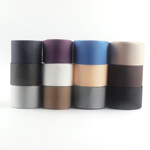 May include: A collection of stacked rolls of woven elastic bands in various colors. The colors include navy, black, silver, brown, purple, white, peach, gray, blue, and dark brown. The bands are tightly woven with a textured surface.