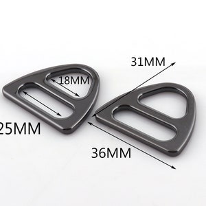 Pode incluir: Dois anéis em D de metal preto com forma triangular. O anel maior mede 36 mm por 31 mm, e o anel menor mede 25 mm por 18 mm.