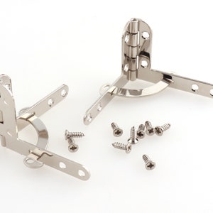 Puede incluir: Dos bisagras de esquina plateadas con un diseño redondeado y un juego de tornillos. Las bisagras son de metal y tienen un acabado pulido.