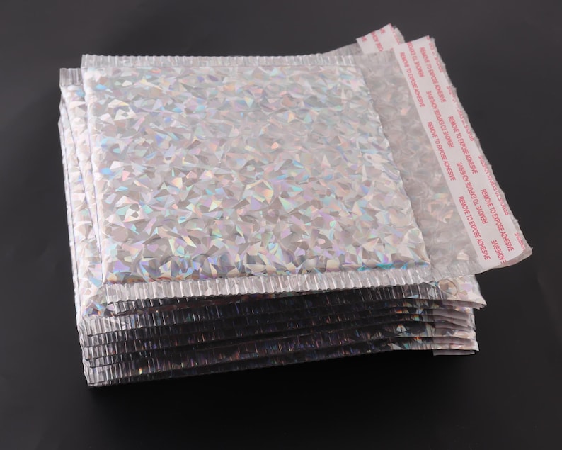 May include: A stack of silver and iridescent bubble mailers with a red adhesive strip that reads "REMOVE TO EXPOSE ADHESIVE".