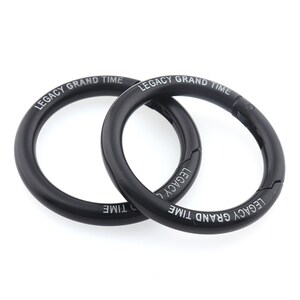 May include: Two black, circular carabiner clips with the text "LEGACY GRAND TIME" printed in white. The clips are made of a smooth, matte material and are designed to open and close with a spring-loaded mechanism.