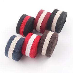 Puede incluir: Surtido de rollos de cinta a rayas de colores. Los rollos presentan varias combinaciones de colores, incluyendo negro con rayas rojas, burdeos con rayas blancas y azul marino con rayas crema. Las cintas están bien enrolladas y parecen estar hechas de un material tejido.