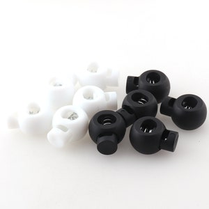 May include: A close-up of a group of white and black plastic toggle clasps. The clasps have a spring mechanism and are used for securing cords or straps.