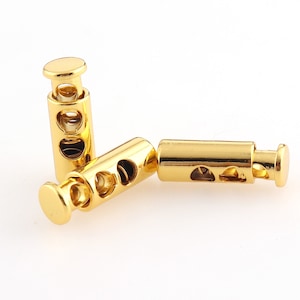 Puede incluir: Tres topes de cordón de metal dorado con un mecanismo deslizante. Cada tope tiene dos agujeros para asegurar los cordones.