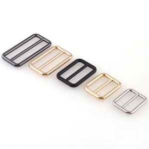 May include: Five metal slider buckles in different sizes and finishes: black, gold, silver, and two shades of gunmetal. The buckles are rectangular with a center bar.