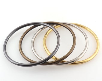 Op de afbeelding: Vier metalen armbanden in verschillende kleuren: donkergrijs, brons, zilver en goud. De armbanden zijn rond en hebben een gladde, gepolijste afwerking. Ze zijn in een rij gerangschikt tegen een witte achtergrond, waarbij hun verschillende metallic tinten worden getoond.