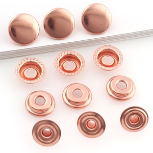 May include: Copper-coloured snap fasteners are displayed. The image shows the top and bottom parts of the snaps. The top pieces are smooth and rounded, while the bottom pieces have a circular design with a central hole.