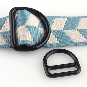 May include: Two black D-rings and a matching slide buckle are displayed against a backdrop of a blue and white patterned strap. The D-rings are made of a glossy black material, and the strap has a geometric design.