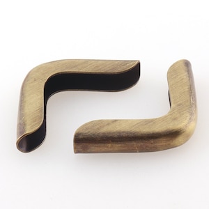 May include: Two antique brass corner protectors, each with a rounded edge and a smooth, slightly textured surface.