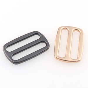 Puede incluir: Dos hebillas deslizantes de metal, una negra y una dorada. Las hebillas son rectangulares con dos ranuras paralelas.