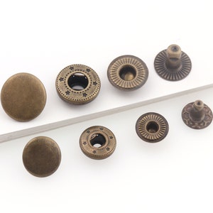 May include: An assortment of antique brass-colored snap fasteners. The collection includes various styles: solid round caps, star-patterned rings, and ridged and patterned backs. These fasteners are ideal for clothing and craft projects.
