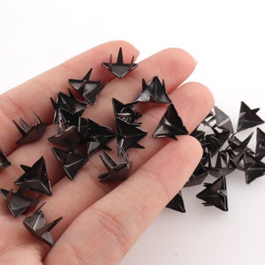 May include: A collection of small, black, triangular metal studs with three pointed prongs. The studs are scattered, with some appearing to be held in a hand. The studs have a shiny, metallic finish and are likely used for decorative purposes.