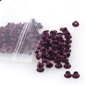 Puede incluir: Una bolsa de ojales metálicos de color morado, con algunos ojales que se caen de la bolsa sobre una superficie blanca.