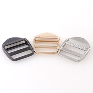 Puede incluir: Tres hebillas deslizantes de metal en negro, dorado y plateado. Cada hebilla tiene una forma rectangular con dos barras horizontales y una parte superior redondeada.