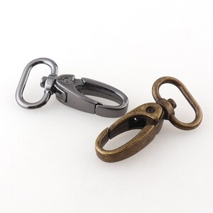 Könnte beinhalten: Zwei Metall-Drehverschlüsse, einer silberfarben und einer antikes Gold. Die Verschlüsse werden zum Befestigen von Riemen oder Ketten an Taschen, Handtaschen oder anderen Gegenständen verwendet.