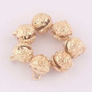 May include: Gold-coloured metal bells with detailed floral patterns. Each bell has a small loop for hanging and a slit for sound. The bells are arranged in a circular pattern, suitable for jewellery making or craft projects.