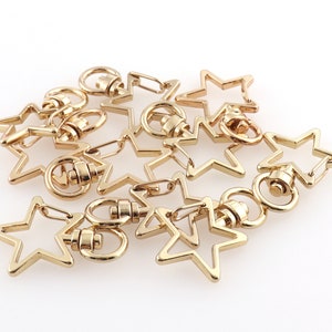 May include: A group of gold-toned metal star-shaped keychains with a swivel clasp. The keychains are arranged in a pile on a white surface.