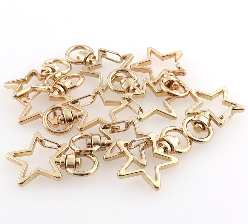 May include: A group of gold-toned metal star-shaped keychains with a swivel clasp. The keychains are arranged in a pile on a white surface.