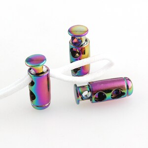 Puede incluir: Tres topes de cordón de metal iridiscente con un cordón blanco enhebrado en dos de ellos. Los topes tienen una forma cilíndrica con una parte superior redonda y un agujero en el centro.