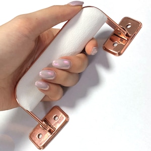 May include: A white leather handle with a rose gold metal frame. The handle is attached to two rose gold metal plates with holes for screws.